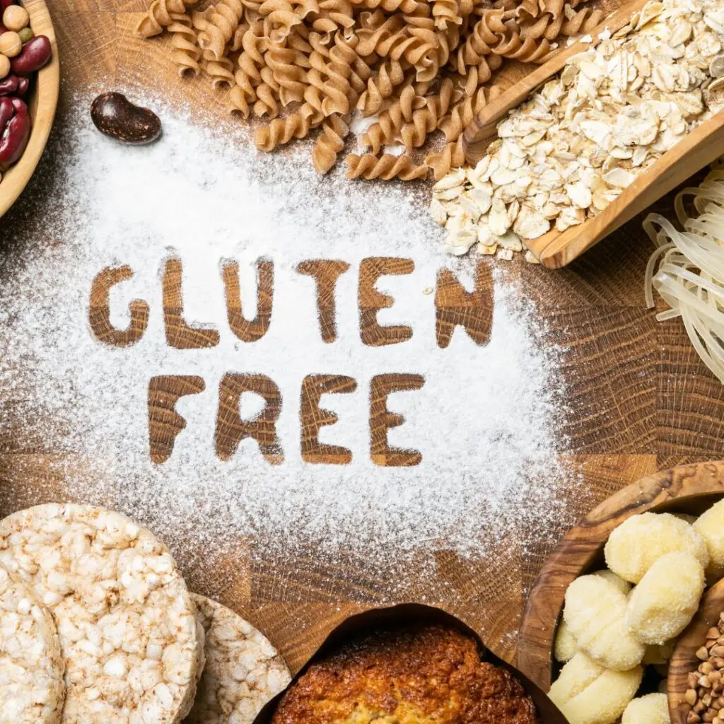 Teksten 'Gluten Free' skrevet i mel omgivet af glutenfri ingredienser såsom pasta, havregryn, riskiks og bønner på en rustik træoverflade