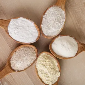 Fem typer glutenfrit mel, herunder majsmel, rismel, mandelmel og havremel, præsenteret i træskeer på en træbordsoverflade
