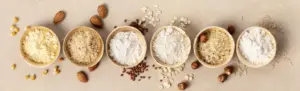 Et udvalg af glutenfri meltyper, herunder kikærtemel, mandelmel, ris- og havremel, arrangeret i små skåle med nødder og kerner på en neutral baggrund.